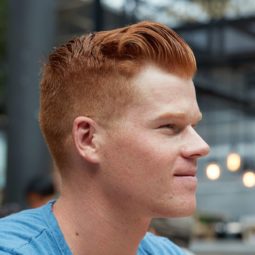 Hair styling products for men: Side shot of a man with short ginger hair styled into quiff with an undercut, wearing blue top and posing in a outdoor setting.