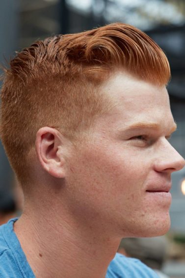 Hair styling products for men: Side shot of a man with short ginger hair styled into quiff with an undercut, wearing blue top and posing in a outdoor setting.