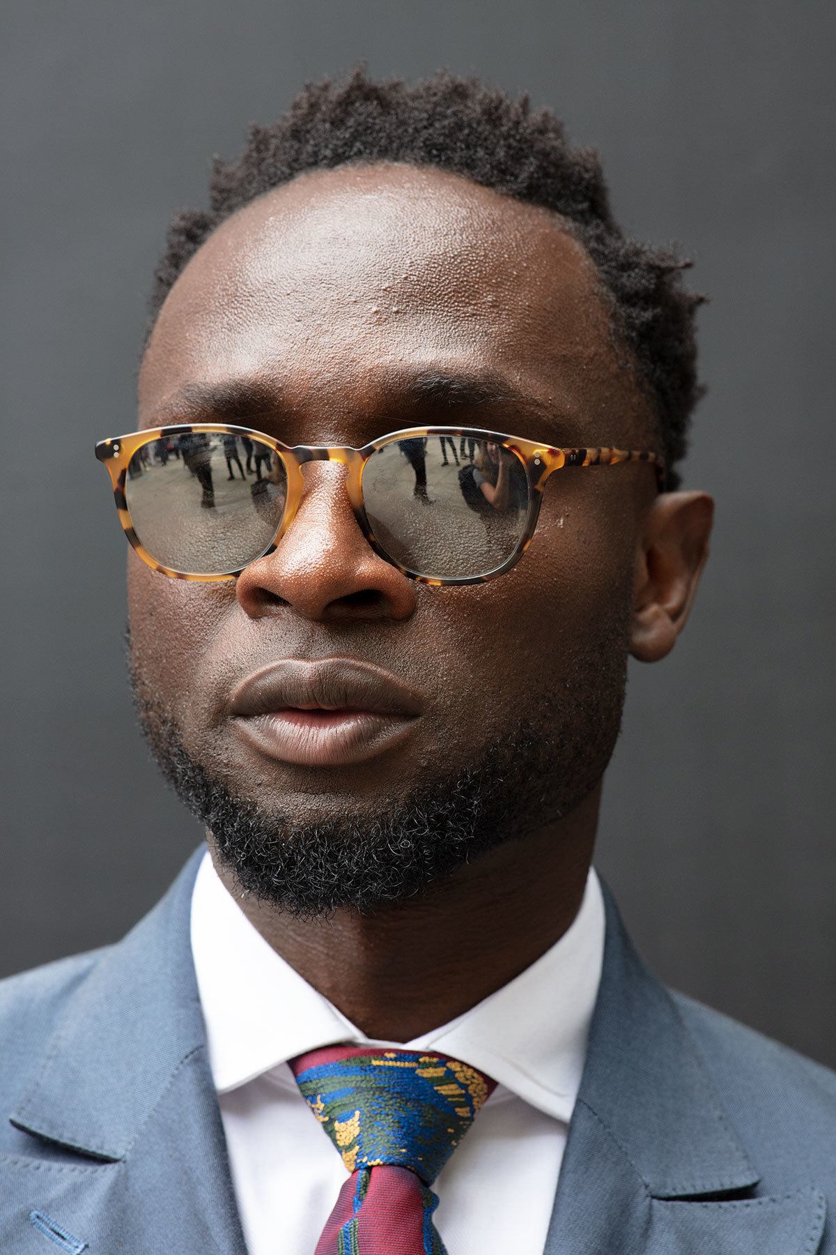 Best hair styling products for men: Close up shot of a man with short dark brown afro hair, wearing sunglasses with a suit and posing outside.