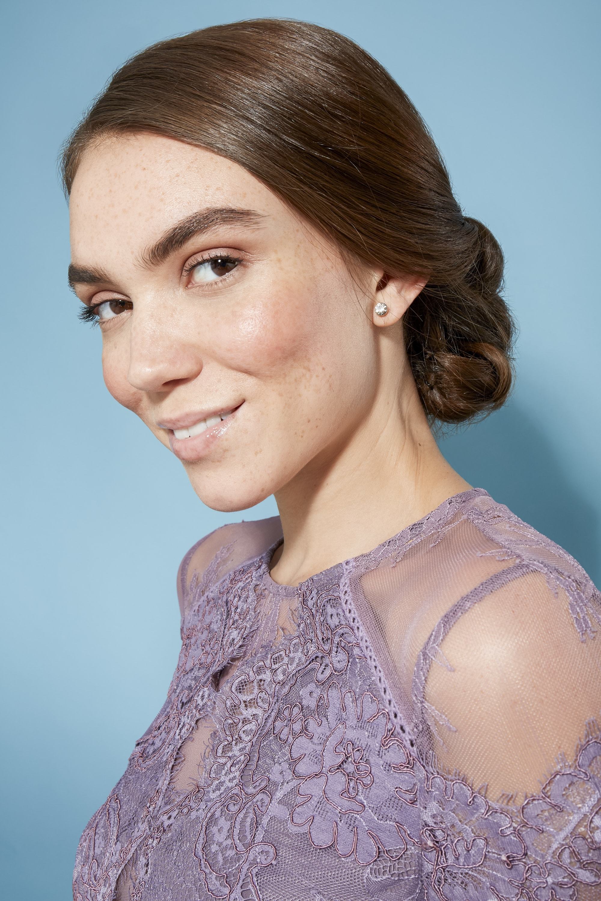 model with brown straight hair styled ina side chignon updo wearing light purple sher and lace top