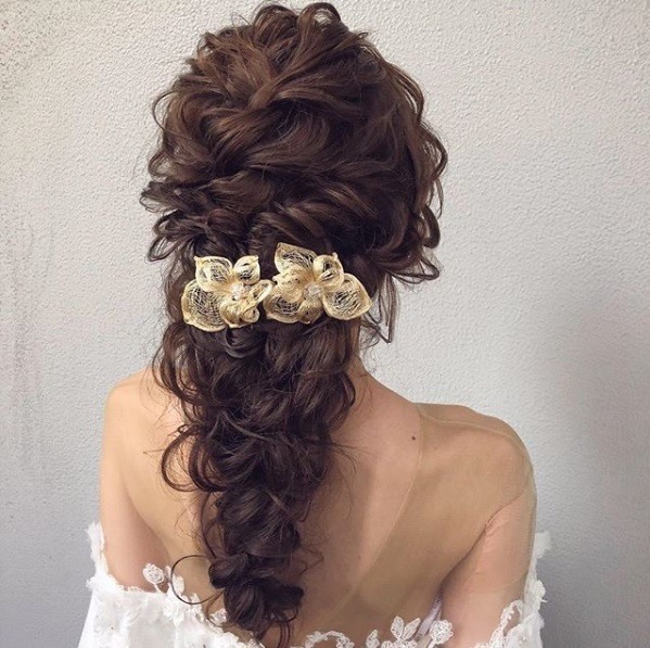 Wedding hairstyles for long hair: Back view of a woman with warm-toned brown curly hair in a twisted braid with floral accessories