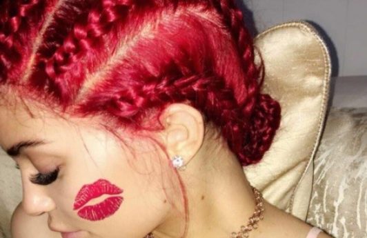 Kylie Jenner's red hair with cornrows posing to the side with a kiss of red lipstick on her cheek