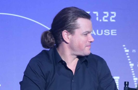 image of matt damon sat down holding a microphone with his long brown hair worn in a low man bun