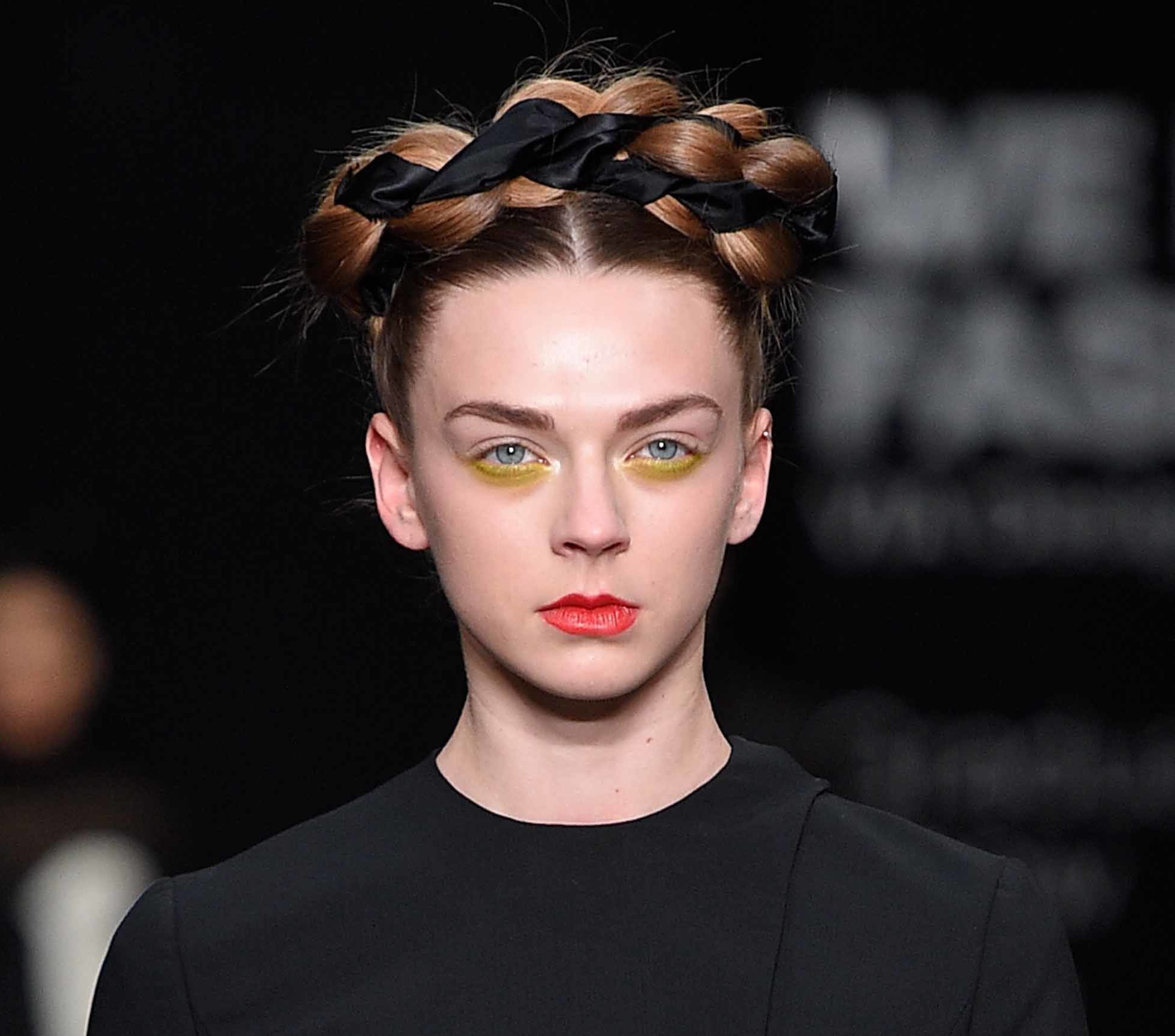 model with brown auburn hair with crown braid woven with ribbon wearing yellow eye makeup, red lips and black dress at aw16 fashion week