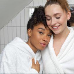 Hair oil treatment guide: Shot of two woman, one with curly hair, the other with straight hair, wearing bath robes in a bathroom setting
