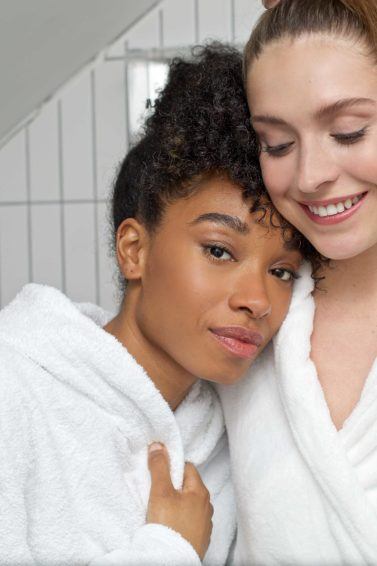 Hair oil treatment guide: Shot of two woman, one with curly hair, the other with straight hair, wearing bath robes in a bathroom setting