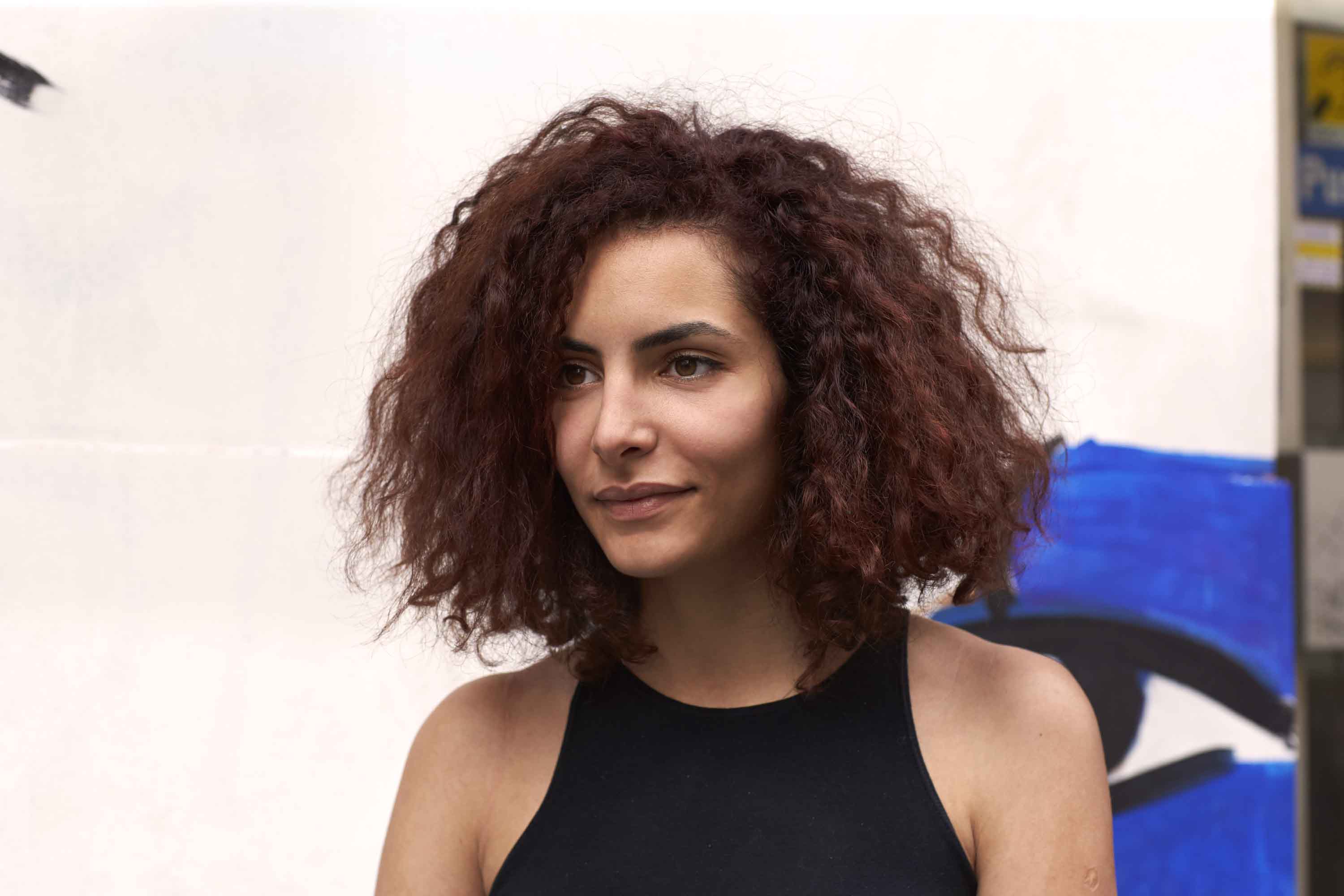 Medium hairstyles for thick hair: Street style shot of a woman with mid-length red brown naturally curly hair, wearing a black top