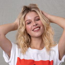 How to make hair look thicker: Blonde model with wavy shoulder length hair with her hand in her hair, wearing a white and red t-shirt