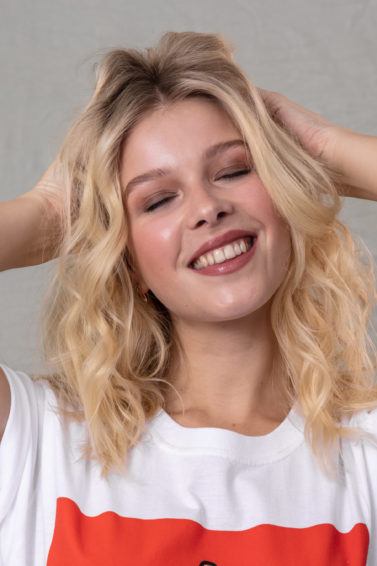 How to make hair look thicker: Blonde model with wavy shoulder length hair with her hand in her hair, wearing a white and red t-shirt