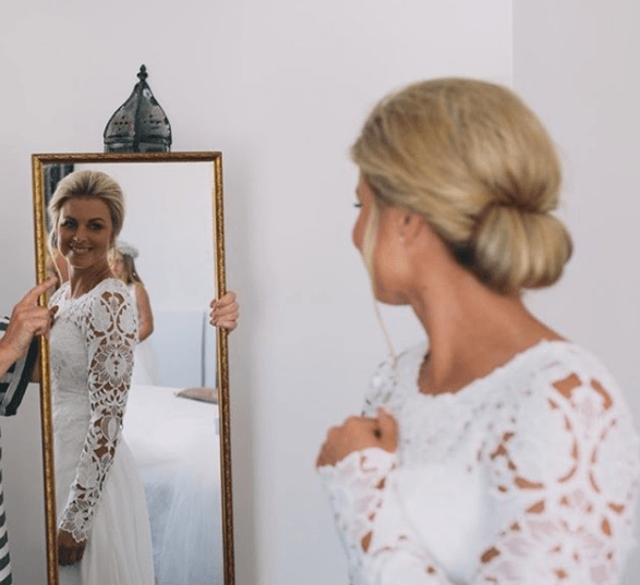 Gibson girl hair: Image of a woman in a wedding dress looking in the mirror with blonde hair and a gibson tuck hairstyle