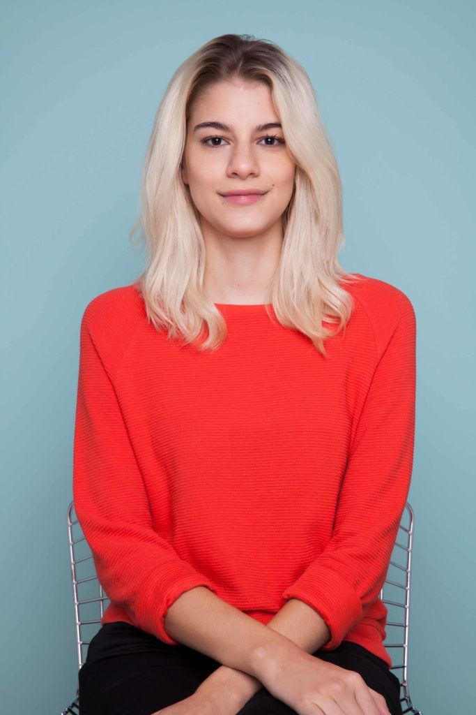 snake braid hairstyles: Blonde woman with medium length hair sitting in studio with blue background wearing a red jumper.