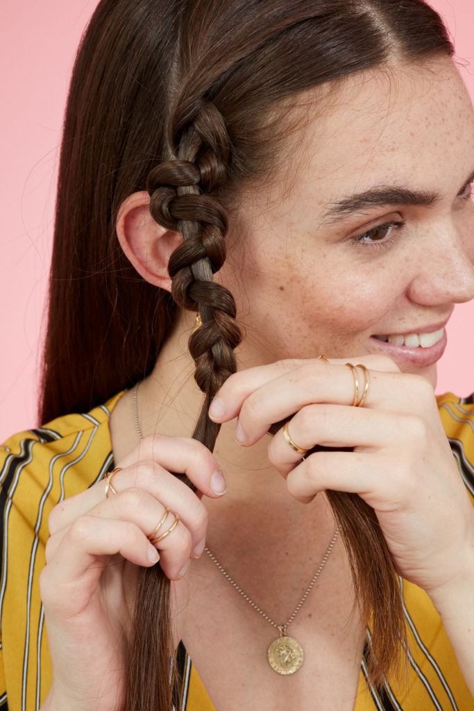 Snake braid hairstyles:Brunette model with long straight hair creating a snake braid. Picture taken in a studio with pink background. Model is wearing a yellow striped dress.