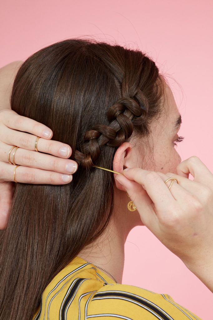 Snake braid hairstyles:Brunette model with long straight hair securing a snake braid behind her ear using bobby pins. Picture taken in a studio with pink background. Model is wearing a yellow striped dress.