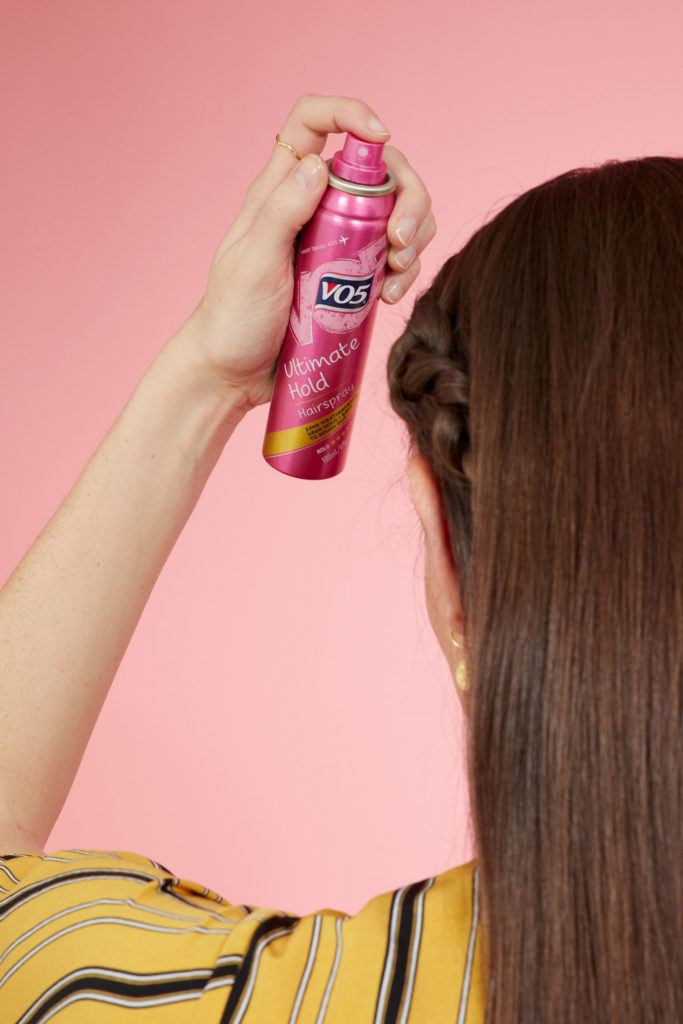 Snake braid hairstyles: Back view of a brunette model with long straight hair spraying hairspray. Picture taken in a studio with pink background. Model is wearing a yellow striped dress.