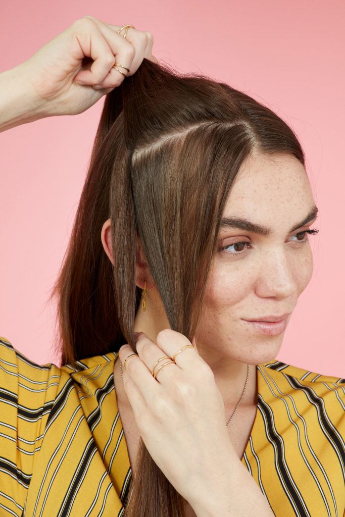 Snake braid hairstyles: Brunette model with long straight hair creating a parting from her crown to ear. Picture taken in a studio with pink background. Model is wearing a yellow striped dress.