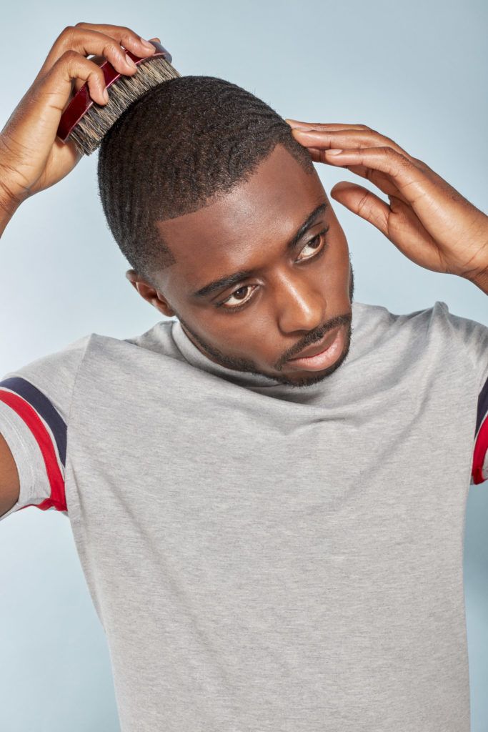 360 waves: Man with short afro hair worn in 360 waves with facial hair wearing a grey t-shirt, using a brush to brush the back of his hair