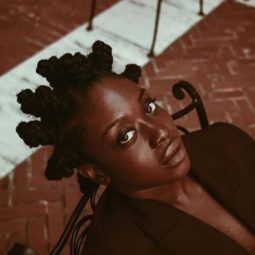 Black history month hair: Close up shot of woman with dark brown hair styled into bantu knots, wearing brown blazer in a living room setting