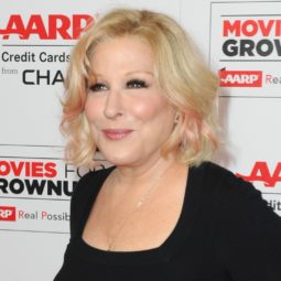Bette Midler on the red carpet wearing black top and navy spriped trousers with her hair worn in a blonde bob cut with pink highlights