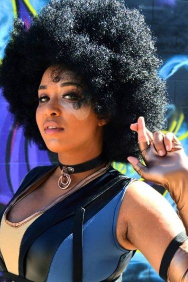 Black hair ideas for Halloween: Woman dressed as Domino from Deadpool 2, with a big dark brown afro, wearing a costume and crossing her fingers