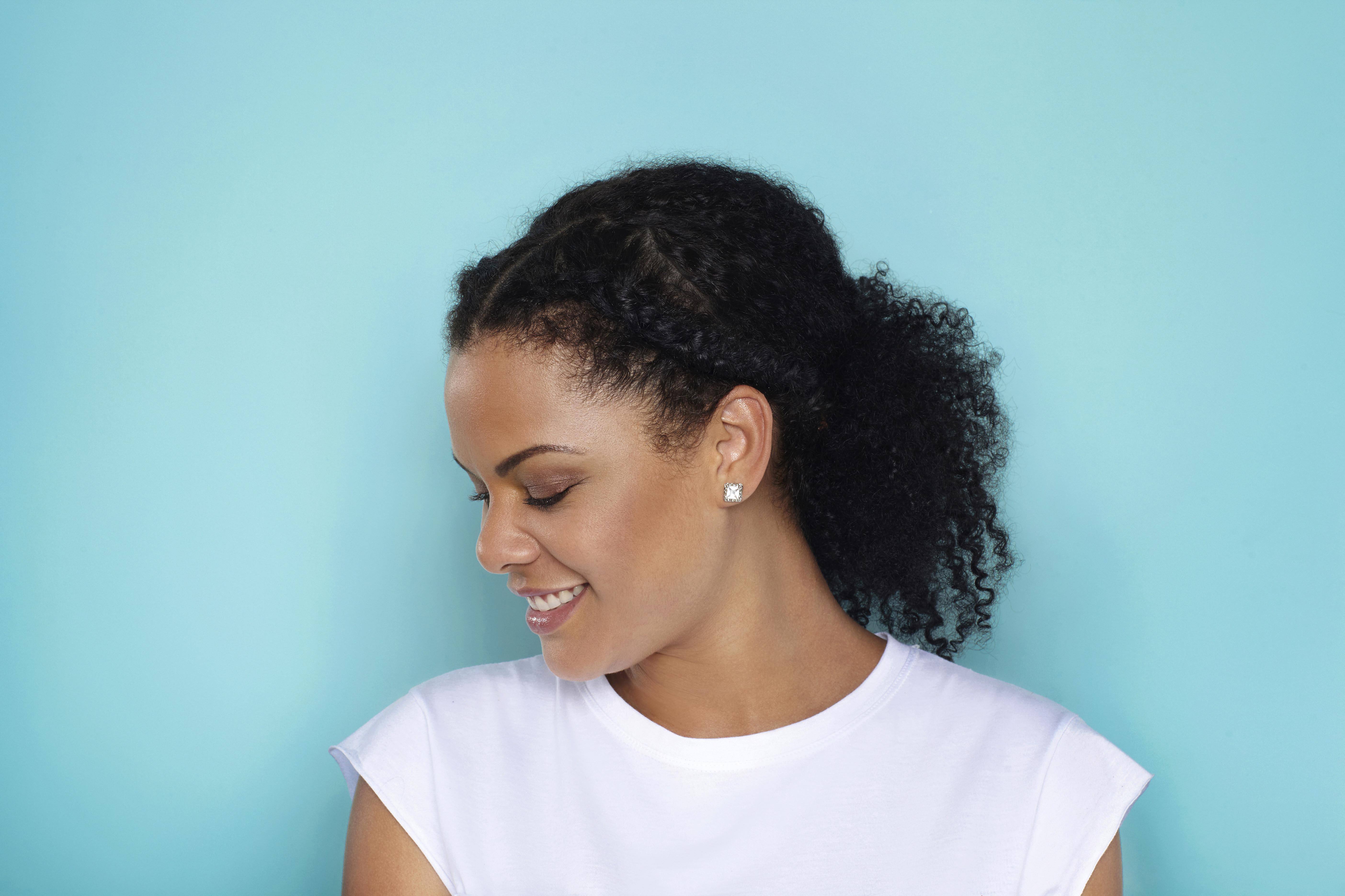 Curly hair hairstyles: side profile of woman with naturally curly hair in ponytail with braids at the front of the hair standing against a light blue backdrop wearing a white tshirt.