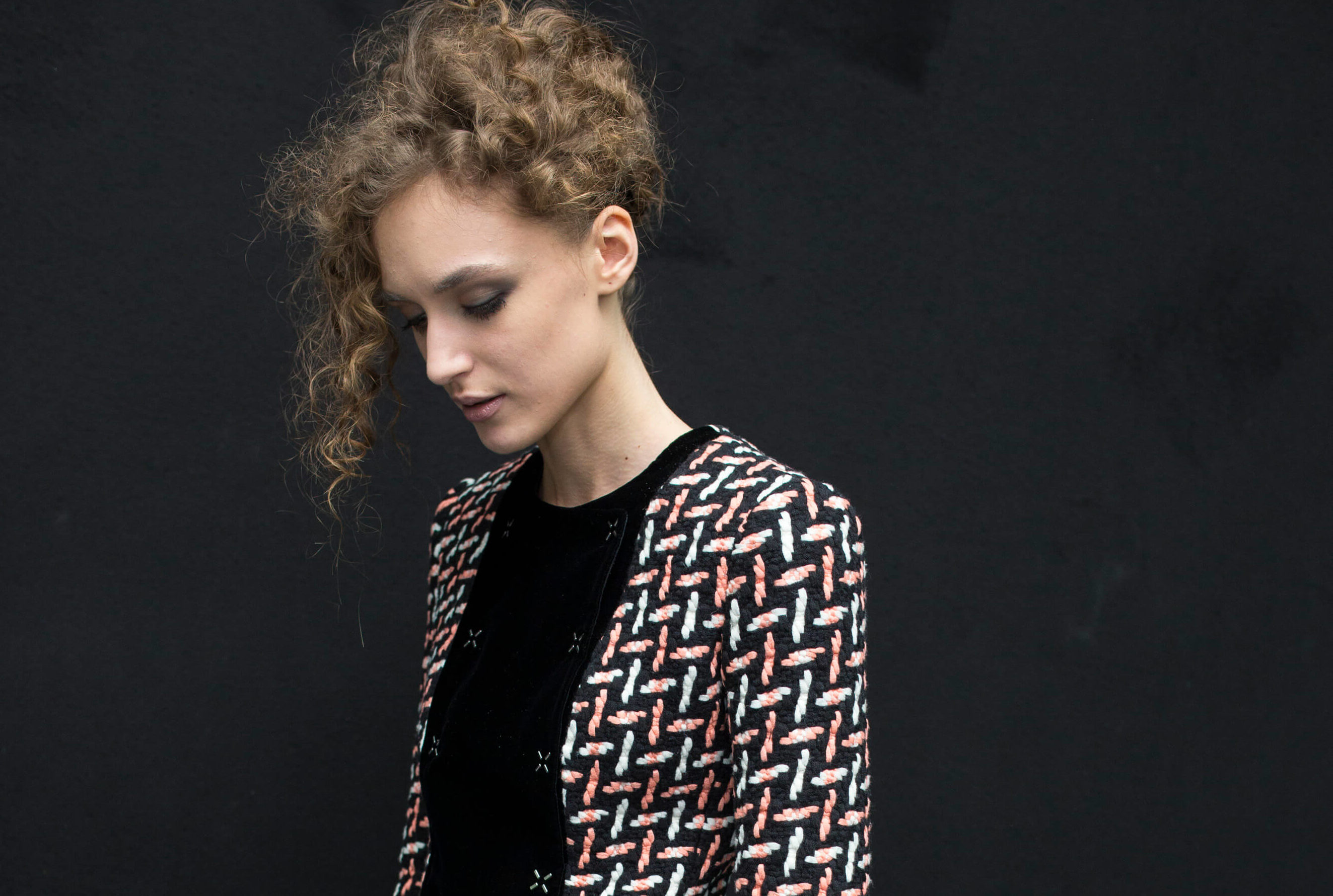 Curly hair hairstyles: woman with curly hair swept up in '80s style standing against a black backdrop wearing pattern jacket