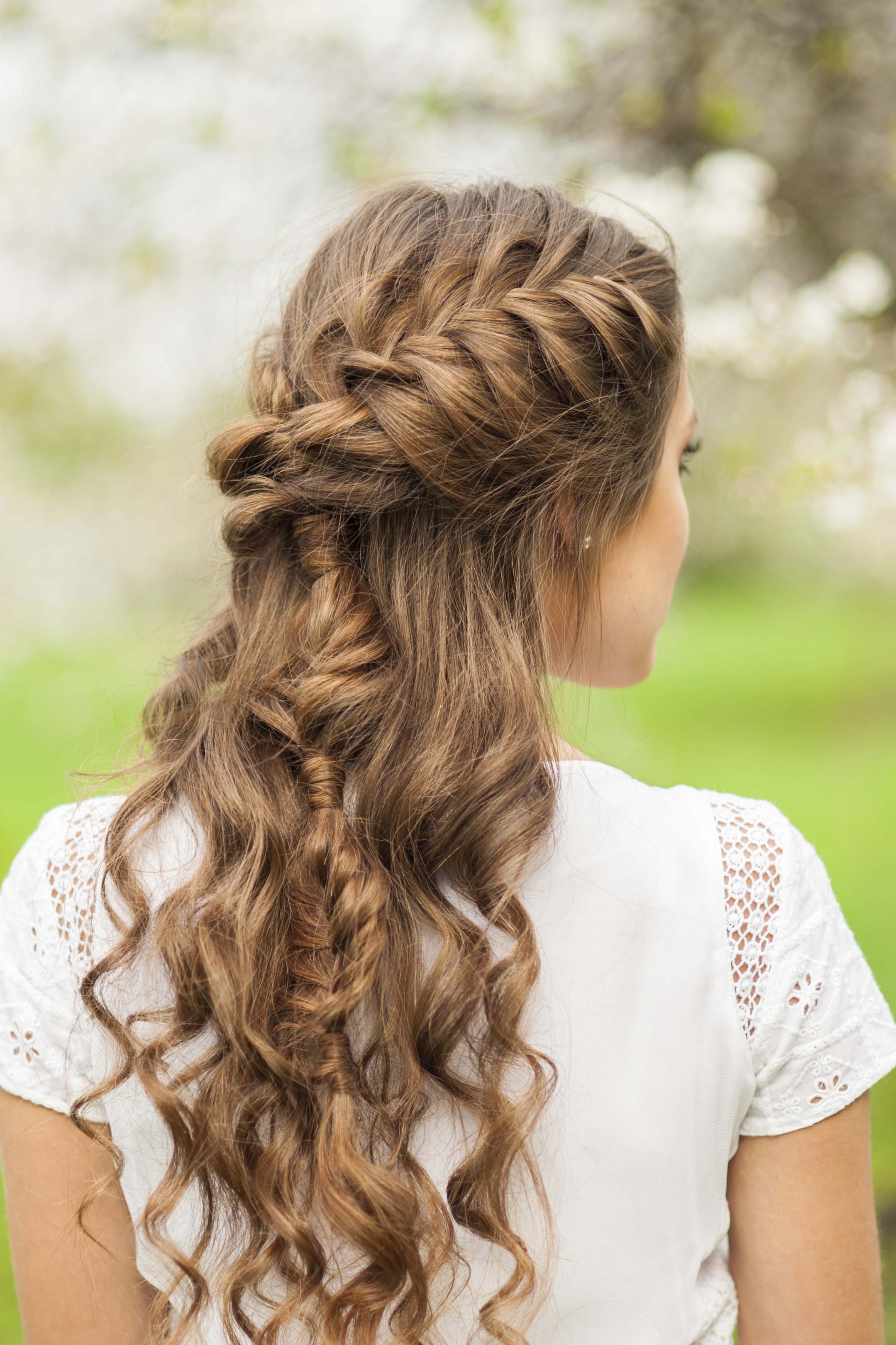Curly hair hairstyles: back view of woman with light brown curly hair with hair in french braid half-up style in outdoor photoshoot
