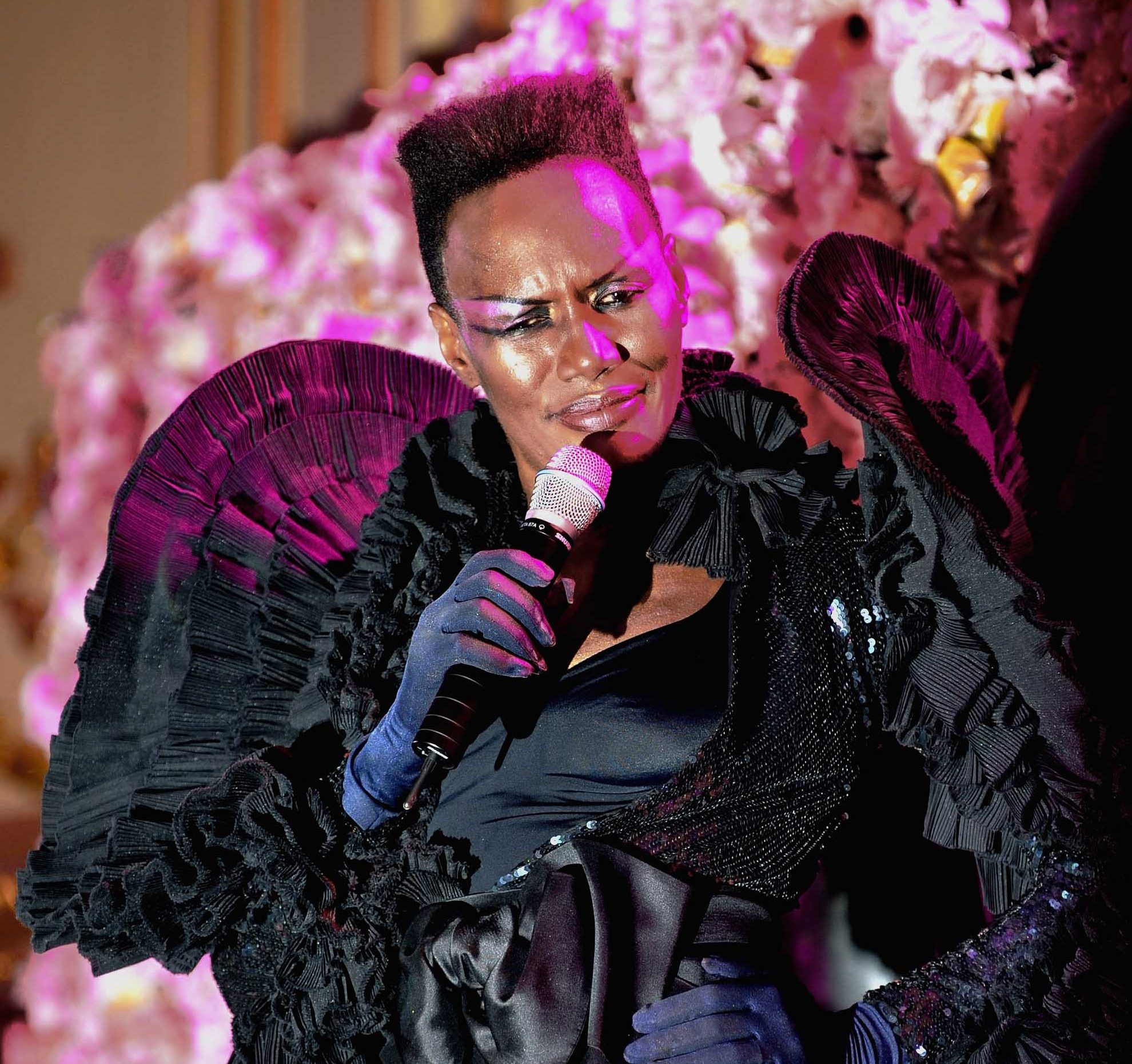 Black history month hairstyles: Close up shot of Grace Jones with her signature short, afro high top hairstyle, wearing all black and posing on the stage