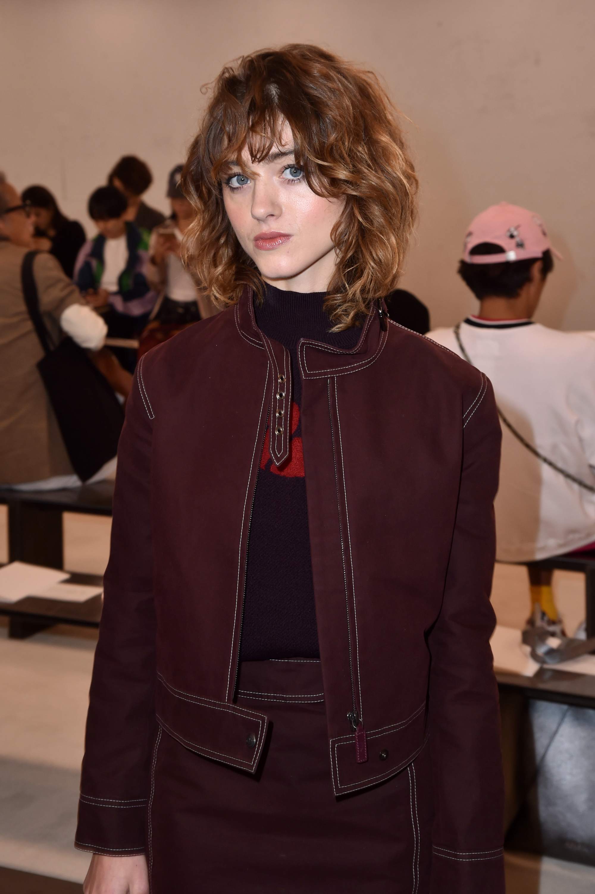 Natalia Dyer with shoulder-length shaggy chestnut brown hair with shaggy fringe, wearing a red denim jacket and skirt