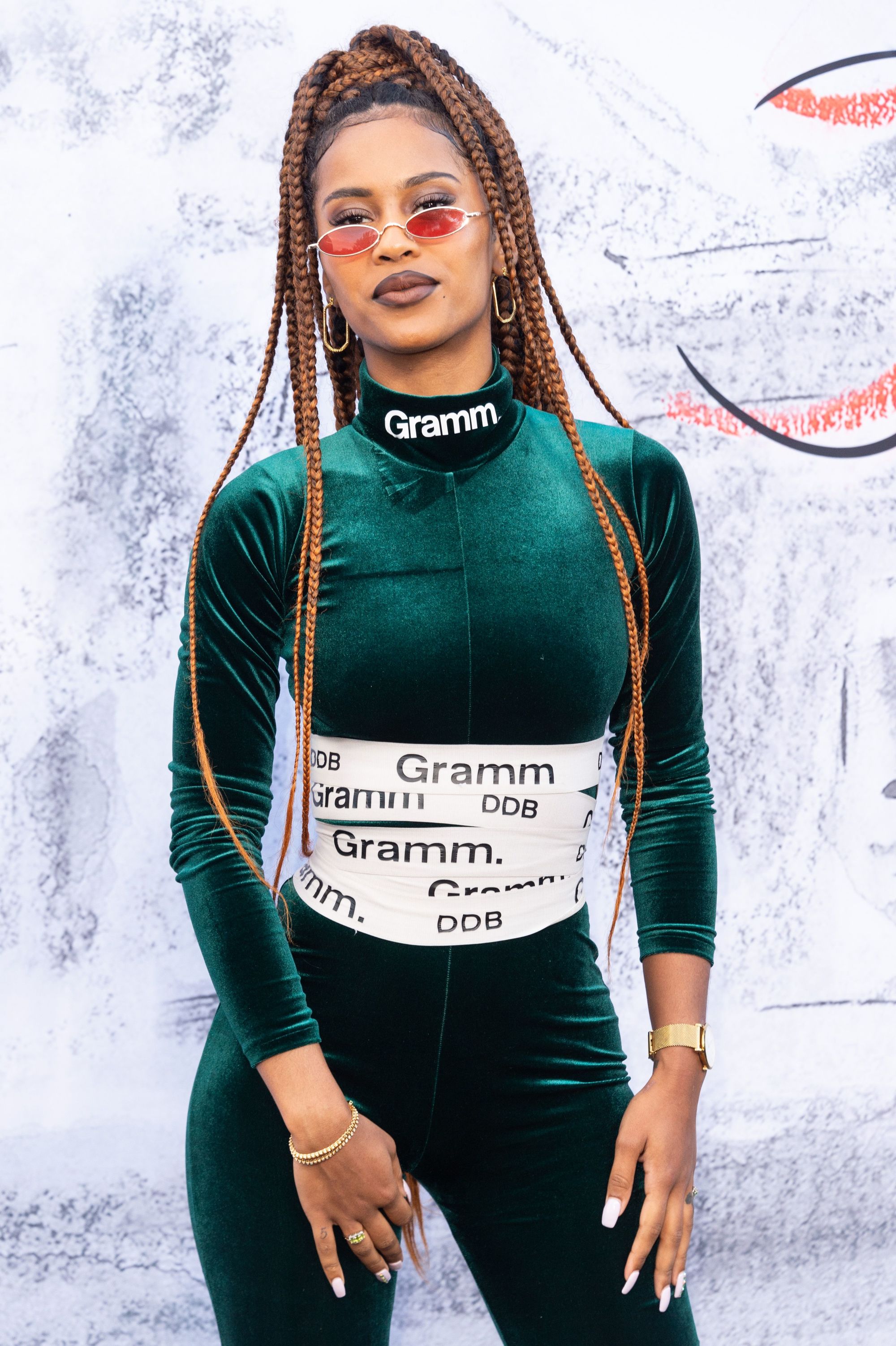 Black history month hairstyles: Close up shot of musical artist IAMDDB with caramel brown dookie braids styled into a high ponytail, wearing a velvet green tracksuit top and bottom