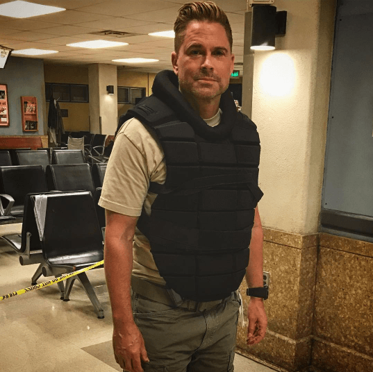 Rob Lowe on set filming wearing a black bullet proof vest and khaki trousers with his hair styled into a quiff