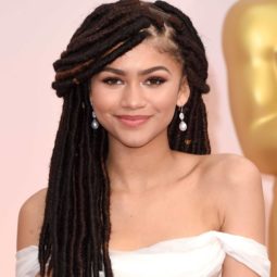 Black history month hairstyles: Close up shot of Zendaya with long dark chocolate faux locs styled into a half-up, half-down style, wearing white bardot dress and posing on the red carpet