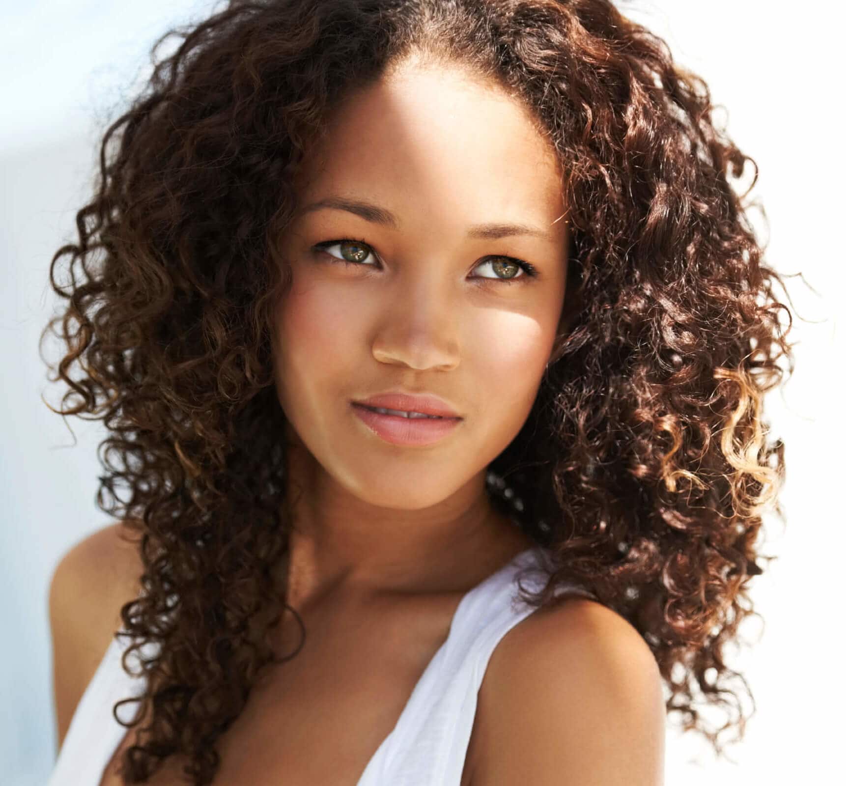 Close up picture of a woman with naturally curly brown hair in twist out curls wearing a white vest.