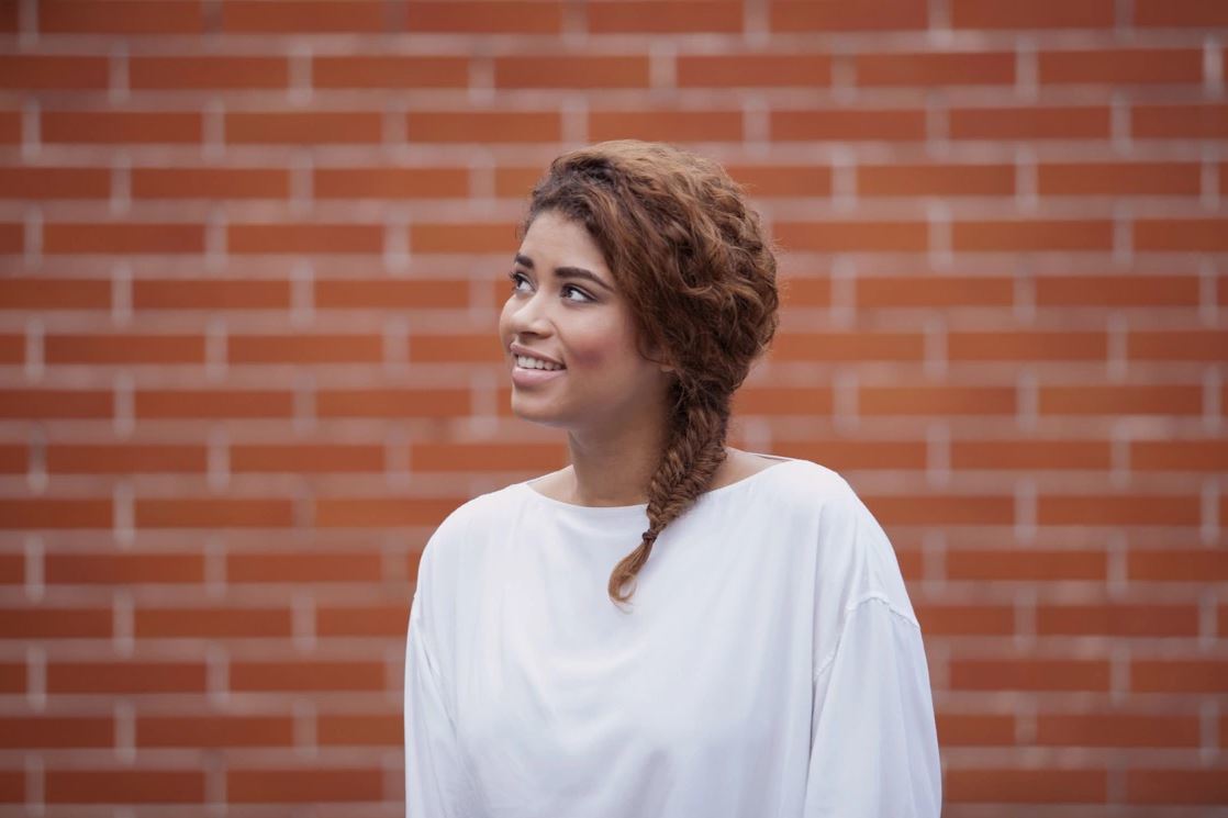 Woman with golden brown naturally curly hair styled in a side fishtail braid wearing a white top standing against a wall backdrop.