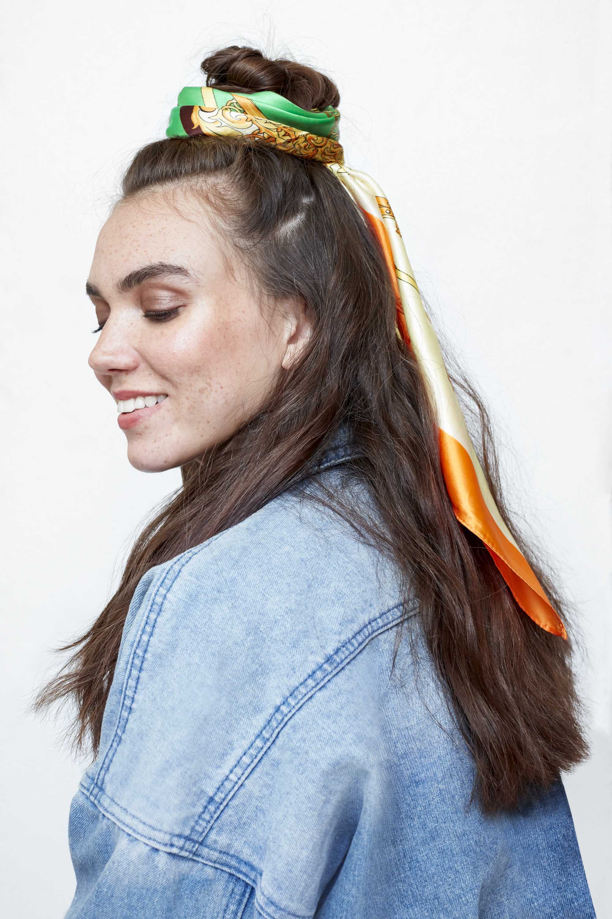 freckled brunette model with her long dark wavy hair in a half up half down bun with a green and orange headscarf tied around the bun wearing a denim jacket