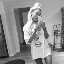 Ariana Grande taking a black and white selfie, wearing a long t-shirt dress with a logo on it, wearing her hair up in a half up bun