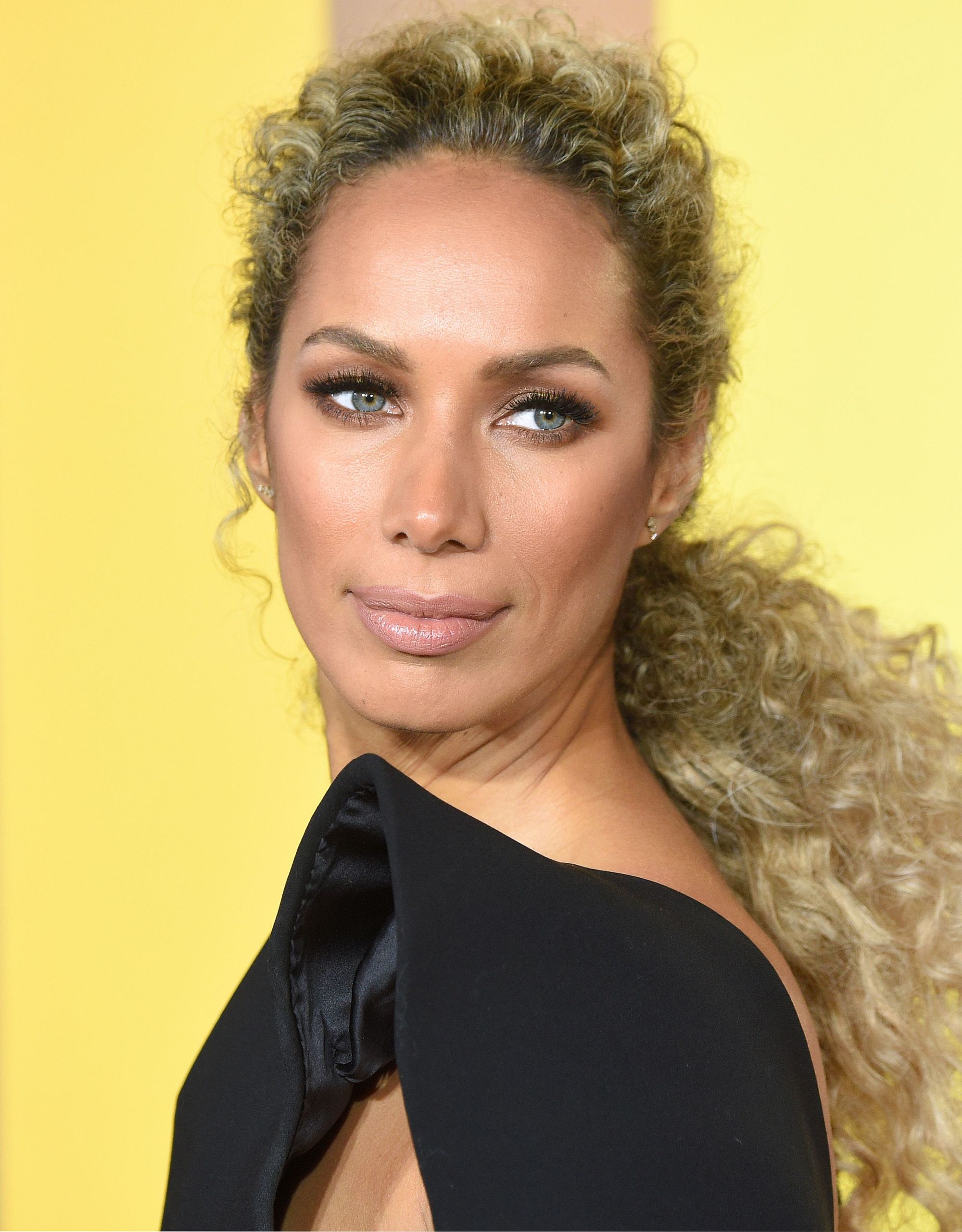 Curly hair hairstyles: Leona Lewis with long golden blonde bronde naturally curly hair styled in a textured low ponytail standing against a yellow backdrop at the Black Panther premiere in London