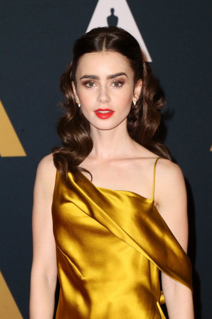 actress lily collins on the red carpet wearing a silk golden gown with her glossy brunette hair worn in a vintage hollywood waves hairstyle