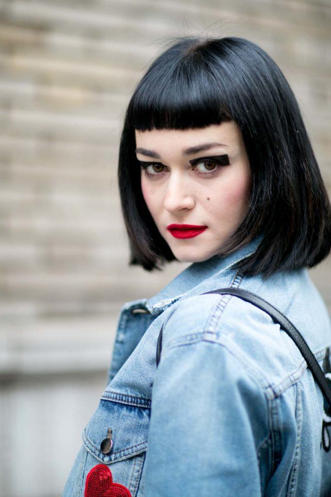 woman wearing a denim jacket and a black backpack with her dyed black hair cut into a blunt bob and short blunt fringe