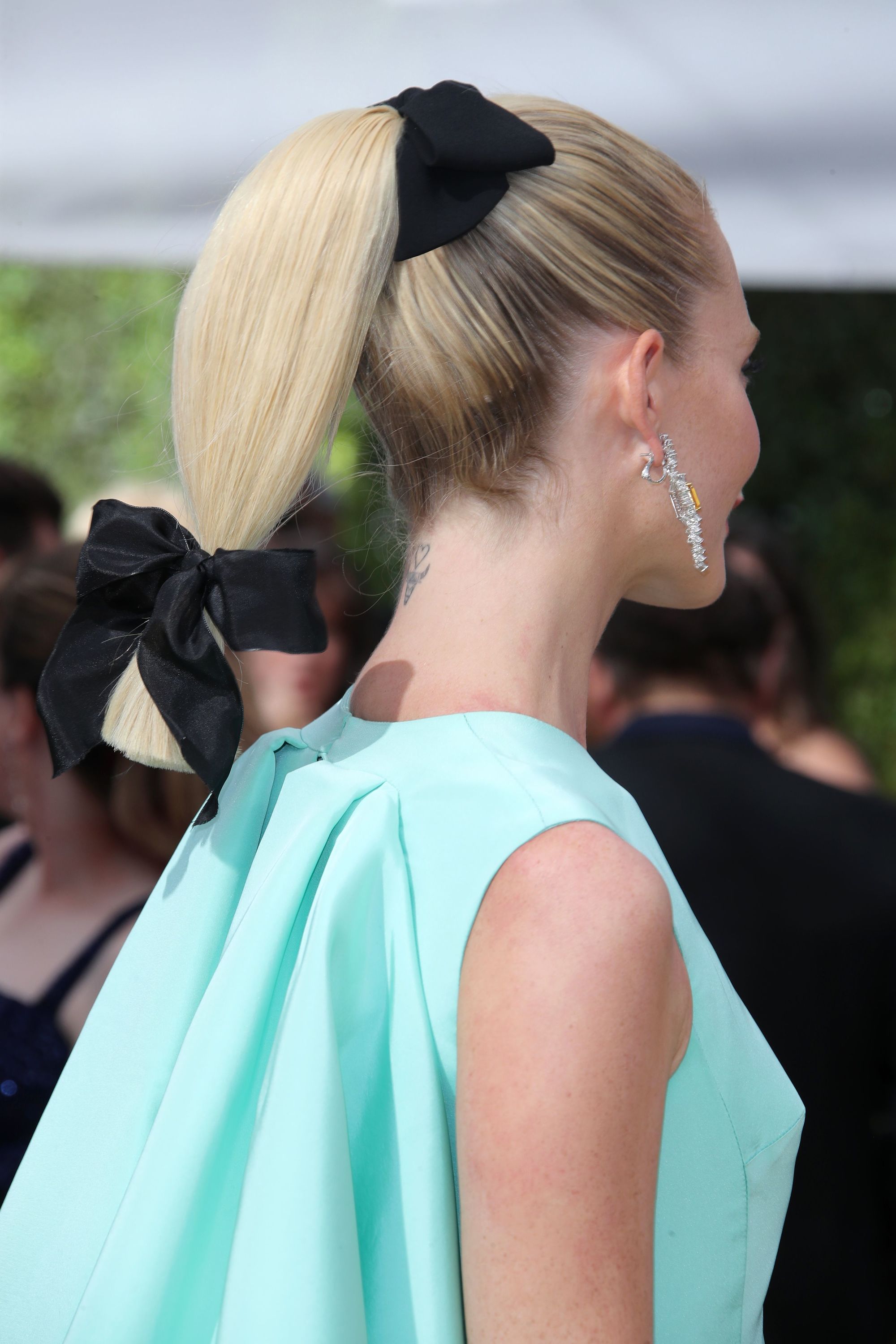 Poppy Delevingne on the red carpet with her medium bleach blonde hair styled into a low sleek ponytail with hair bows at the base and the end of it, wearing a duck blue dress