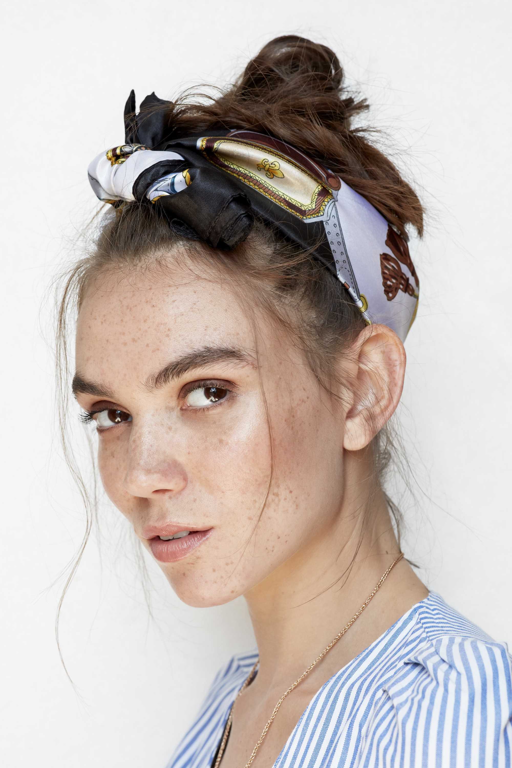 close up head shot of a freckled brunette model with her hair in a messy bun updo with a printed blue and gold scarf wrapped around it tied in a bow
