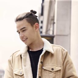 street image of a young man wearing a shearling jacket and black top with his medium brown hair worn up in a top knot with an undercut