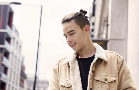 street image of a young man wearing a shearling jacket and black top with his medium brown hair worn up in a top knot with an undercut