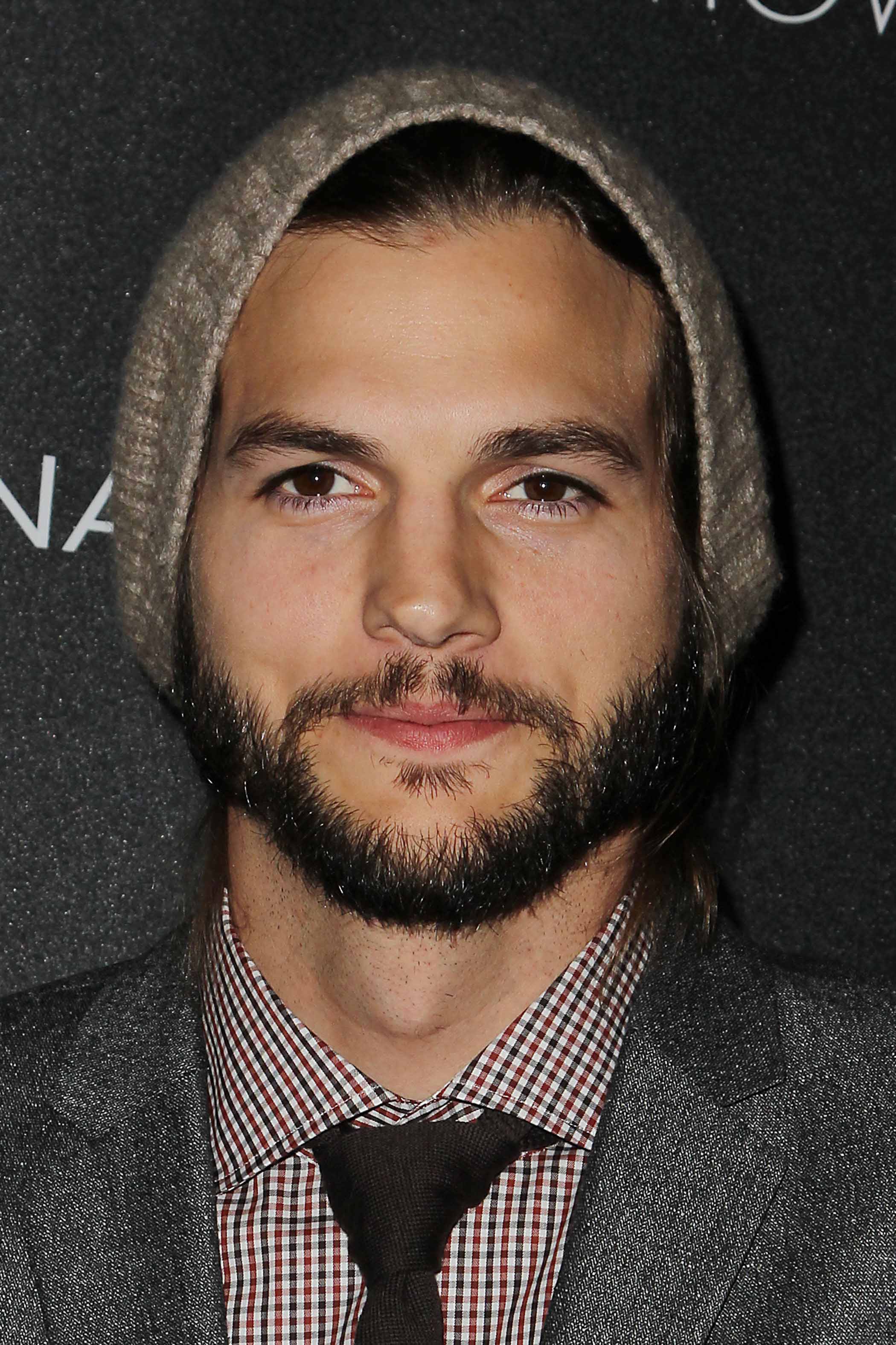 Ashton Kutcher on the red carpet wearing a grey suit and tie with a beanie hat on and neat facial hair