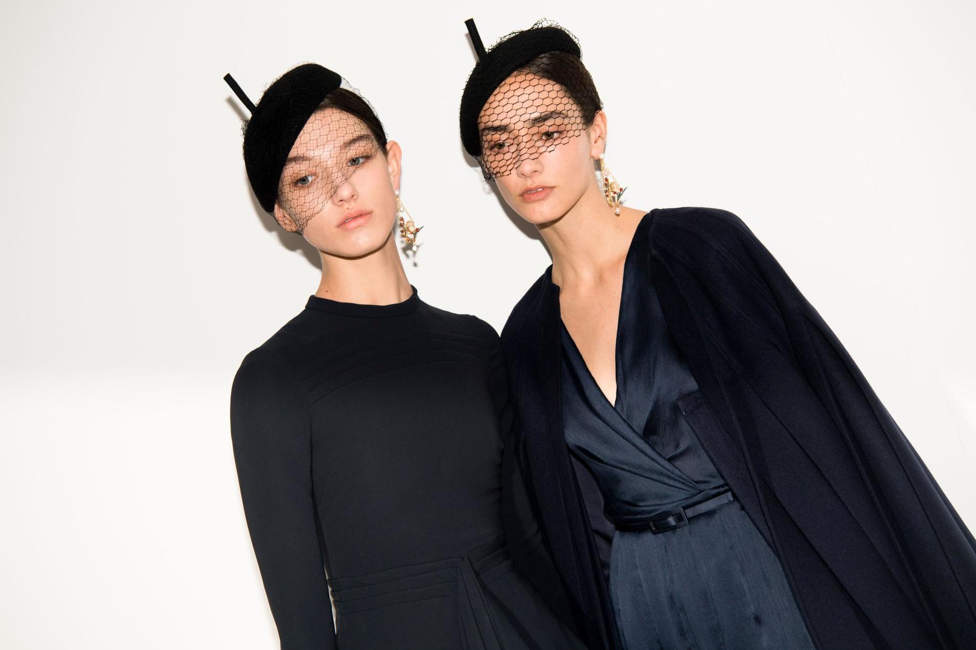 Retro hairstyles: Close up shot of two women with sleek low buns topped with pillar box/beret style hats with net, backstage at Christian Dior