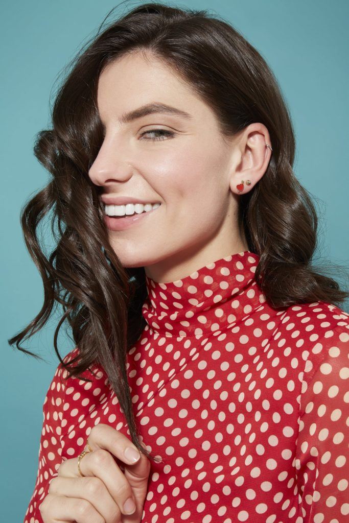 woman with dark brown glossy hair in loose curls looking at the camera smiling and wearing a red and white spotted dress