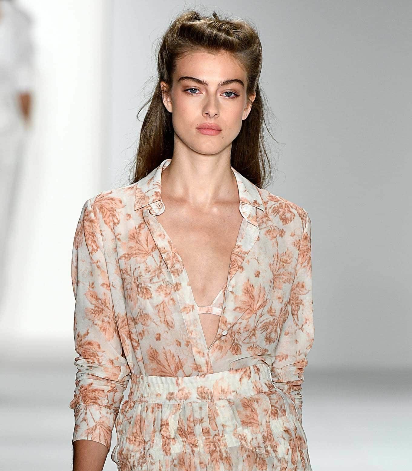 Retro hairstyles: Close up shot of a woman with medium brown long hair styled into a modern half-up, half-down rolled updo walking on the runway