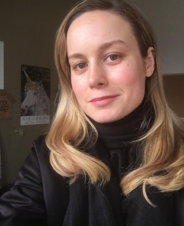 actress Brie Larson with long golden blonde hair worn in loose waves at the ends wearing an all black outfit in a room with a unicorn poster in the background