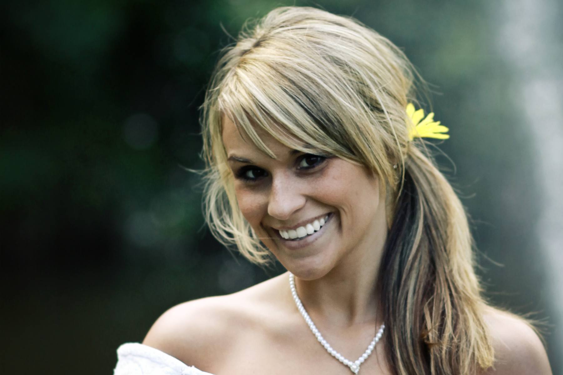 blonde bride wearing a white dress and necklace with her hair in a side ponytail with a flower hair accessory