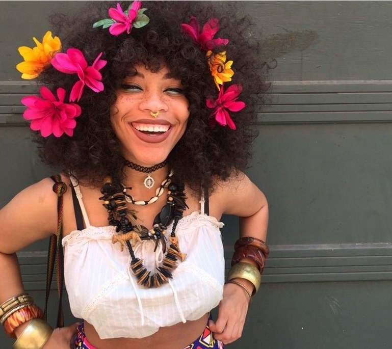 close up shot of woman with big curly afro hair flower hair accessories in it, wearing white crop top and bangles and posing outside