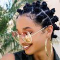 natural hairstyles for short hair: close up shot of woman with short natural hair styled into small bantu knots, wearing sunglasses and big hoop earring, wearing denim and posing outside